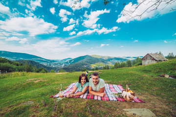 Fototapeta premium Couple in love on a white and red plaid in a field on a picnic. against a background of a mountains. romantic moment. Man and woman with champagne glass.
