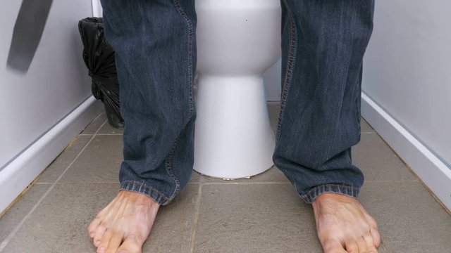 The Legs of a Man Sitting and Stand up from a Public Toilet. A man in jeans and bare feet in a white toilet cubicle sits down on the toilet. Defecation process. Toilet in a public place.