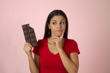 attractive and happy hispanic woman in red top thinking and doubting holding chocolate bar on pink...