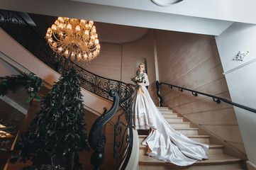 A beautiful bride in a lace dress with a long train is standing on the steps in a room with a beautiful interior.