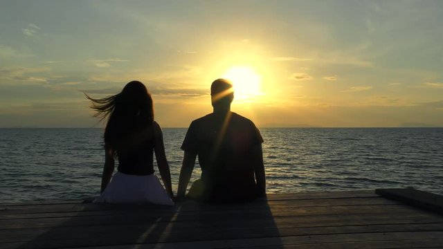 Romantic couple Sitting on the Pier at colorful sunset on background. Young Couple at the Sea of Tropical Island