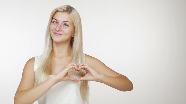 slowmotion caucasian smiling girl in white with long blond hair showing heart shape by fingers in good mood isolated over white background. Concept of emotions