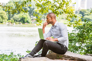 Fototapeta premium Young American hispanic man wearing gray shirt, black pants, leather shoes, sitting by lake at Central Park, New York, working on laptop computer, looking up, hand touching chin, thinking..