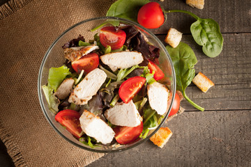 Fresh salad made of tomato, ruccola, chicken breast, arugula, crackers and spices. Caesar salad in a white, transparent bowl on wooden background