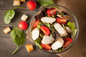 Fresh salad made of tomato, ruccola, chicken breast, arugula, crackers and spices. Caesar salad in a white, transparent bowl on wooden background