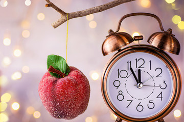 Vintage copper alarm clock showing five minutes to midnight. New Year countdown. Sugar coated red apple ornament hanging on branch. Glittering garland lights. Pastel Colors. Christmas greeting card