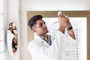 Handsome ophthalmologist cleaning eyeglasses frame in optical store. 