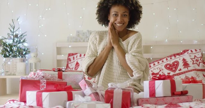 Excited young woman with a pile of Christmas gifts clapping her hands in delight as she celebrates the holiday season at home