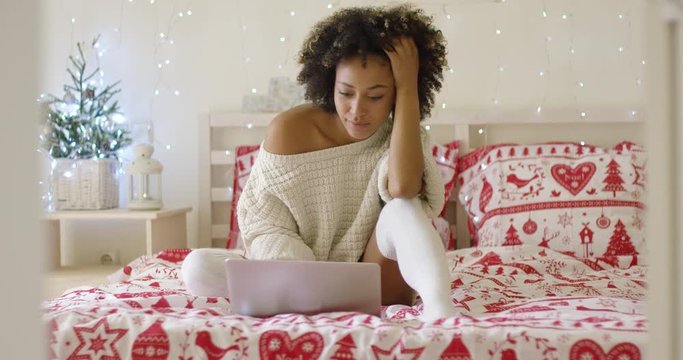 Serious beautiful woman in oversized sweater and long white stockings seated on bed with Christmas theme decorations using laptop computer