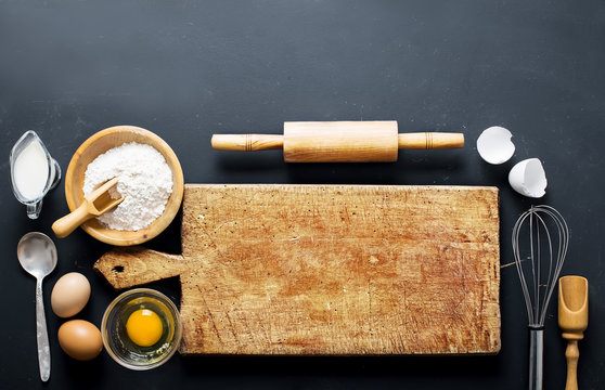 Baking Ingredients. Bowl, Eggs, Flour, Eggbeater, Rolling Pin And Eggshells On Black Chalkboard From Above.