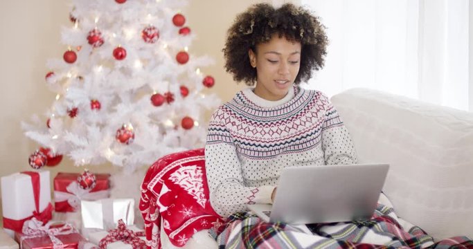 Happy excited young woman reading a Christmas message on her laptop computer as she relaxes on a sofa in front of the tree and gifts