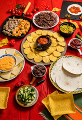 Mexican food concept: tortilla chips, guacamole, salsa, chilli, refried black beans, pulled beef, chicken, cheese and fresh ingredients over vintage red rustic wooden background. Top view
