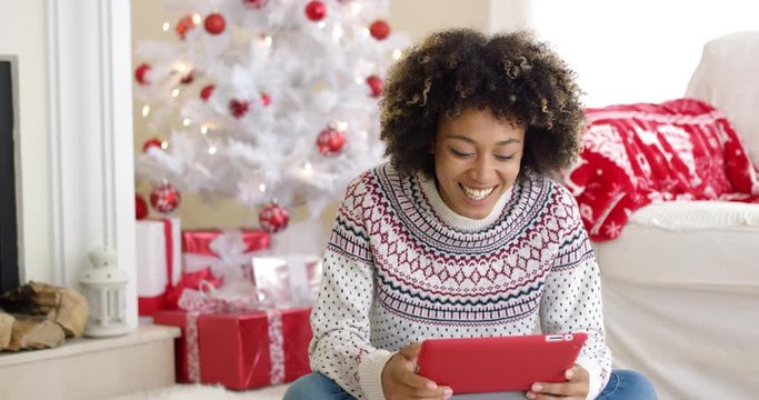 Young woman surfing the internet at Christmas sitting on the floor using a tablet pc in front of the decorated tree and piles of gifts