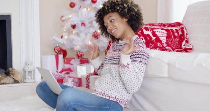 Young woman having a video chat on her laptop waving at the computer as she sits in a red and white themed Christmas living room