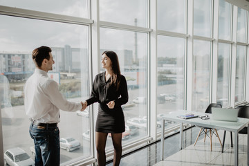Businessman and businesswoman shaking hands together while standing in front of office building windows overlooking the city