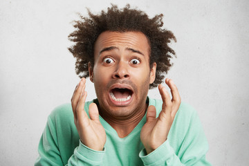 Portrait of dark skinned mixed race Afro American male looks with surprsied expression, has crisp hair, being scared to hear horrified news, isolated over white background. Facial expression