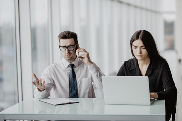 Young creative team brainstorming in the office. Handsome male businessman making call while smiling assistant sitting next to him and working on laptop.
