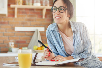 Smiling delightful young female wears casual clothes and spectacles, makes list of household...