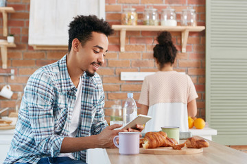 Fototapeta na wymiar Black man in casual wear checks email or reads world news on electronic device, drinks morning coffee and croissants, sits at wooden kitchen table and his wife in background busy with making salad