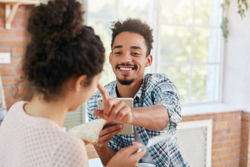 Family couple makes fun at kitchen during lunch: bearded man touches nose of girlfriend who makes sandwiches, feels great love and sympathy. Hipster male uses tablet computer has breakfast with wife