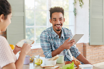 Pleased stylish man has beard and mustache, smiles happily, shares his success at work to wife who prepares supper. Attractive dark skinned male with broad smile reads news on website uses internet