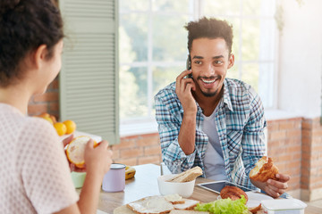 Portrait of joyful man being glad hear old best friend over mobile phone, eats croissant and looks at wife who peels orange. Businessman in casul clothes being at home, arranges meeting with partners