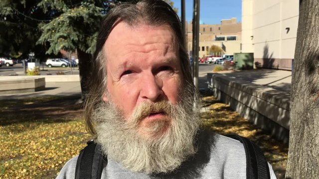 Bearded homeless man speaks towards camera under bright sunlight in downtown setting.  
