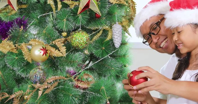 Happy toothless little girl and her father decorating a Christmas tree together while wearing Santa hat at home, shot in 4k resolution