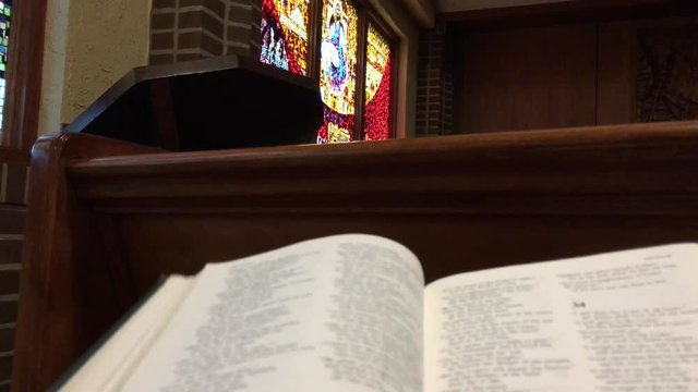 From first person perspective, hand opens up bible in church pew with stained glass windows in background.  