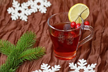 A cup of tea with slice of lemon, little branch of red rosehip fruit, brown background, white snowflakes and green spruce branch