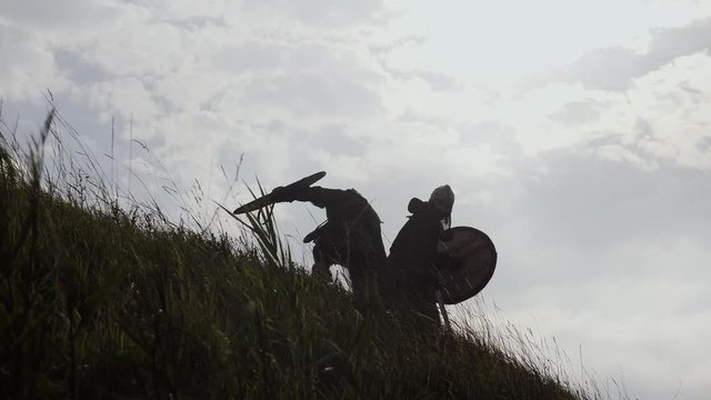 Silhouettes of two warriors Viking are fighting with swords and shields.