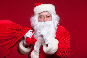 Portrait of Santa Claus with huge red sack keeping forefinger by his mouth and looking at camera