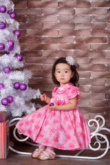 Asian pin up little cute girl wearing pink casual dress posing close to new year christmas white tree with violet purple balls toys and colourful presents