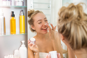 Obraz na płótnie Canvas Young beautiful woman applying moisturising cream on skin in bathroom. Standing in towel with little jar of moisturizer, looking in the mirror, laughing and having fun. Morning skincare routine. 
