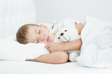 Sick boy with thermometer in mouth lying in bed and hugging a toy bear