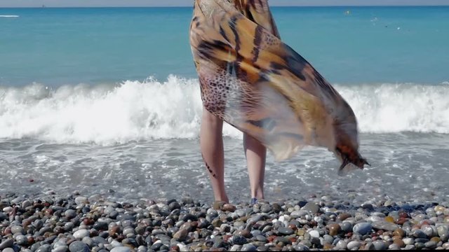Female feet are on a pebble beach. Pareos develops in the wind