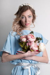 Beautiful young blonde woman with clean skin and flower wreath in her hair on grey background with bouquet of flowers