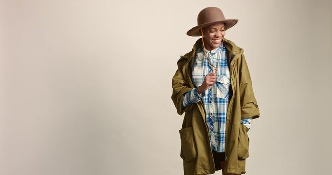 Pretty smiling mixed race female model wearing a plaid shirt over a short black skirt, khaki parka and a brown felt bush hat