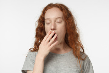 Sleepy redhead girl yawning, waking up, closing her mouth with hand, isolated on gray background