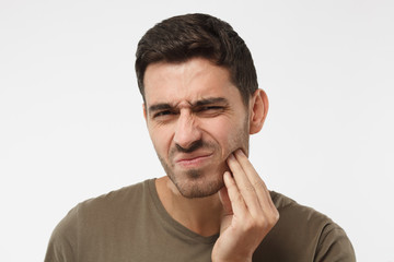 Indoor headshot of young male feeling pain, holding his cheek with hand, suffering from bad toothache, looking at camera with painful expression. Tooth ache concept.