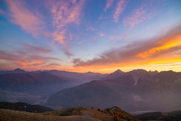 The Italian French Alps at sunset. Colorful sky over the majestic mountain peaks, dry barren terrain and green valleys. Sunburst and backlight expansive view from above.