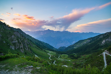 Fototapeta premium Mountain road leading to high mountain pass on the Italian Alps.. Expasive view at sunset, colorful dramatic sky, adventure road trip in summer.