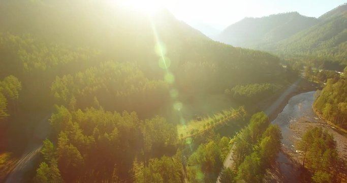 Aerial UHD 4K view. Mid-air flight over fresh mountain river and meadow at sunny summer morning. Green trees and sun rays on horizon. Rural dirt road below.