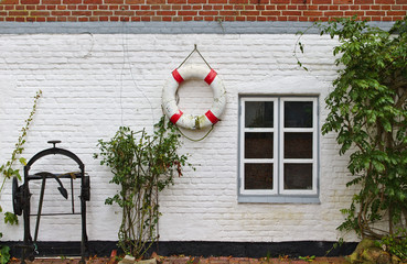 Red and whitewashed brick wall with window, green bushes, red and white life buoy and historic anchor winch