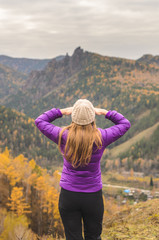 A girl in a lilac jacket looks out into the distance on a mountain, a view of the mountains and an autumnal forest by an overcast day, free space for text.