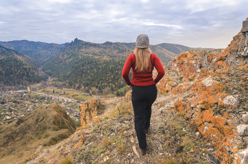 Naklejka na ściany i meble A girl in a red jacket looks out into the distance on a mountain, a view of the mountains and an autumnal forest by an overcast day. Free space for text