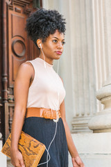 Young black female teacher with afro hairstyle wearing sleeveless light color top, belt, carrying small leather briefcase, walking out from vintage office building, listening music with earphones..