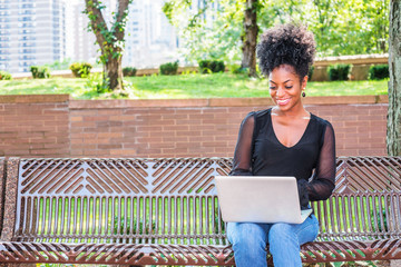 Young beautiful African American Woman with afro hairstyle wearing mesh sheer long sleeve shirt, jeans, sitting on bench at park in New York, looking down, working on laptop computer, smiling..