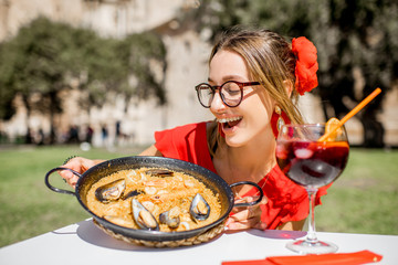 Young woman in red dress with sea Paella, traditional Valencian rice dish, sitting outdoors at the restaurant in the centre of the old town of Valencia