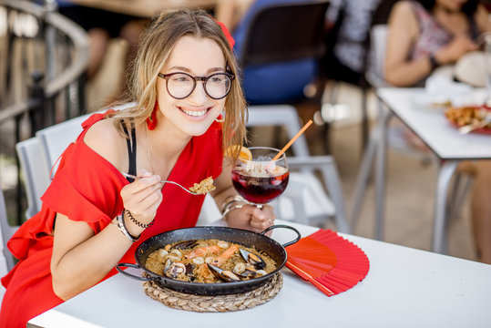 Young woman in red dress eating sea Paella, traditional Valencian rice dish, sitting outdoors at the restaurant in Valencia, Spain